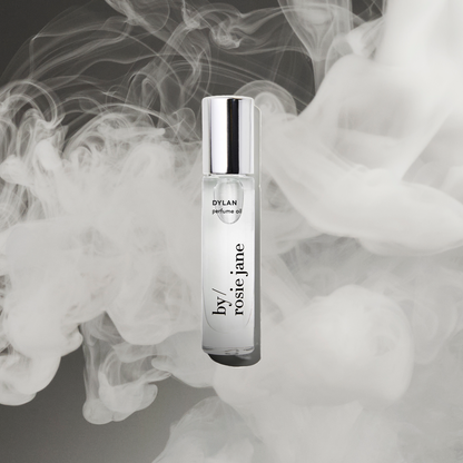 A clear glass DYLAN PERFUME OIL bottle by byrosiejane with a silver cap, centered on a smoky dark gray background with swirling white mist—evoking the allure of white musk and whispers of sexual freedom.