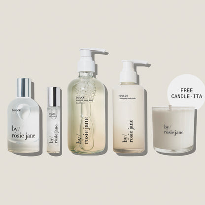 Five By Rosie Jane skincare and body care products arranged on a muted blue-gray background, including a large pump bottle, a small roller bottle, a tall body milk bottle, and a candle jar with a 'FREE CANDLE - ITA' callout. The products are mostly frosted white with clear sections and silver caps.