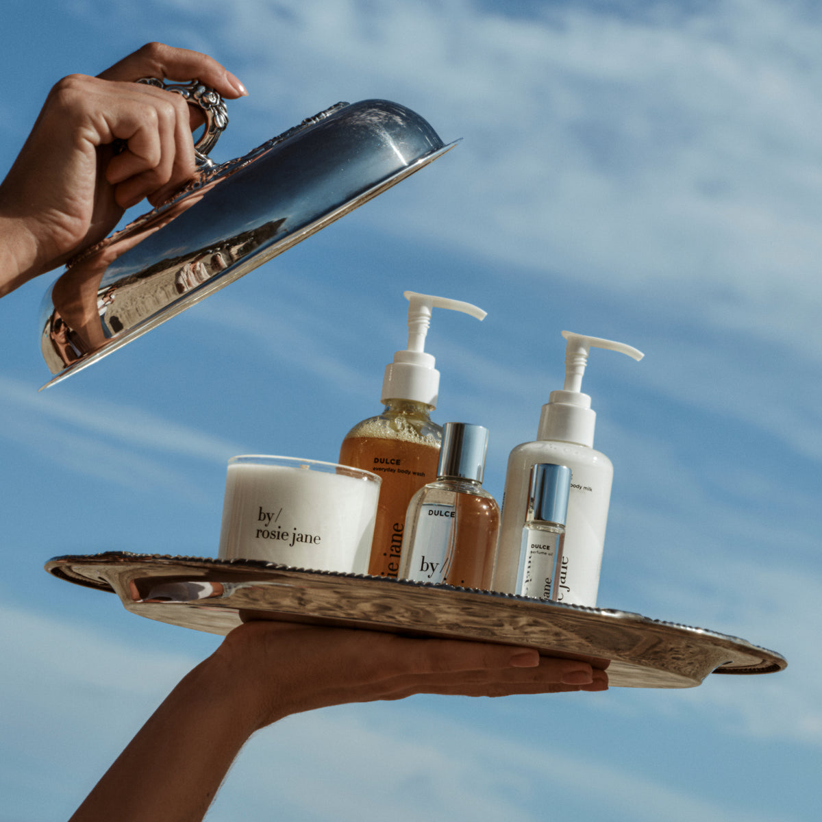 A person holds a silver serving tray outdoors against a blue sky, displaying a curated collection of skincare products from by / rosie june and Dolce & Gabbana. The products include a white candle, a brown liquid soap bottle, a small clear bottle with a silver cap, and a white lotion bottle with a pump dispenser.