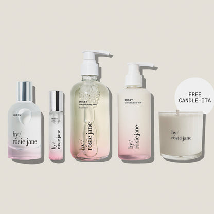 Five By Rosie Jane skincare and body care products arranged on a muted blue-gray background, including a large pump bottle, a small roller bottle, a tall body milk bottle, and a candle jar with a 'FREE CANDLE - ITA' callout. The products are mostly frosted white with clear sections and silver caps.