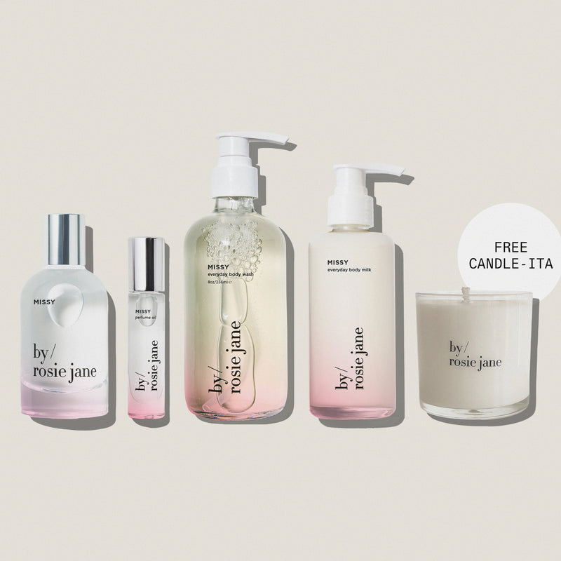 Five By Rosie Jane skincare and body care products arranged on a muted blue-gray background, including a large pump bottle, a small roller bottle, a tall body milk bottle, and a candle jar with a 'FREE CANDLE - ITA' callout. The products are mostly frosted white with clear sections and silver caps.