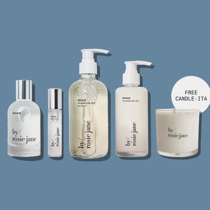 Five By Rosie Jane skincare and body care products arranged on a muted blue-gray background, including a large pump bottle, a small roller bottle, a tall body milk bottle, and a candle jar with a 'FREE CANDLE - ITA' callout. The products are mostly frosted white with clear sections and silver caps.