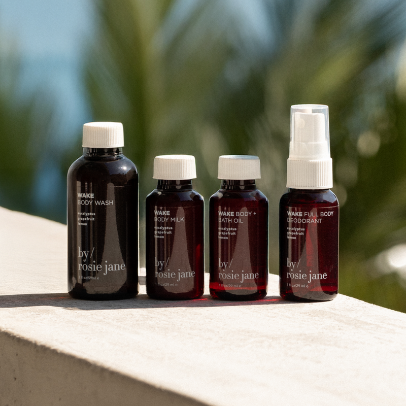 Four bottles of wake 'by rosie jane' skincare products on a ledge with a blurred natural background.