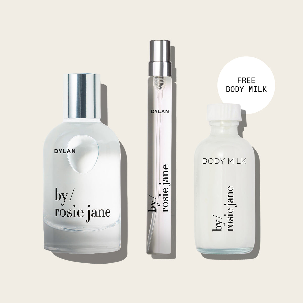 Three Dylan by Rosie Jane fragrance bottles arranged on a light beige background: a large frosted bottle with a silver cap, a slim matching spray vial, and a frosted bottle labeled BODY MILK. A white circular callout says FREE BODY MILK.