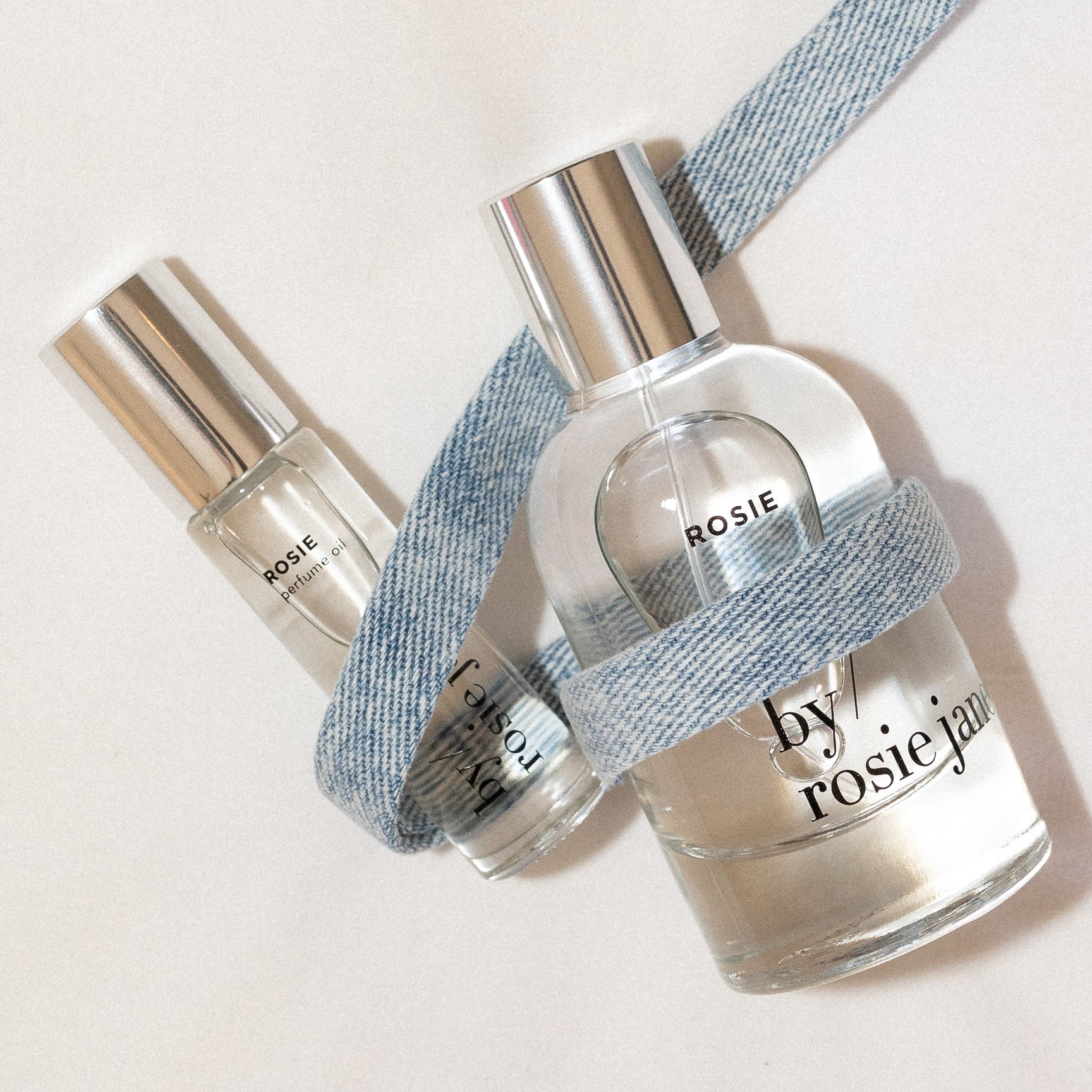 Two 'by rosie janie' perfume bottles with denim ribbon on a light background