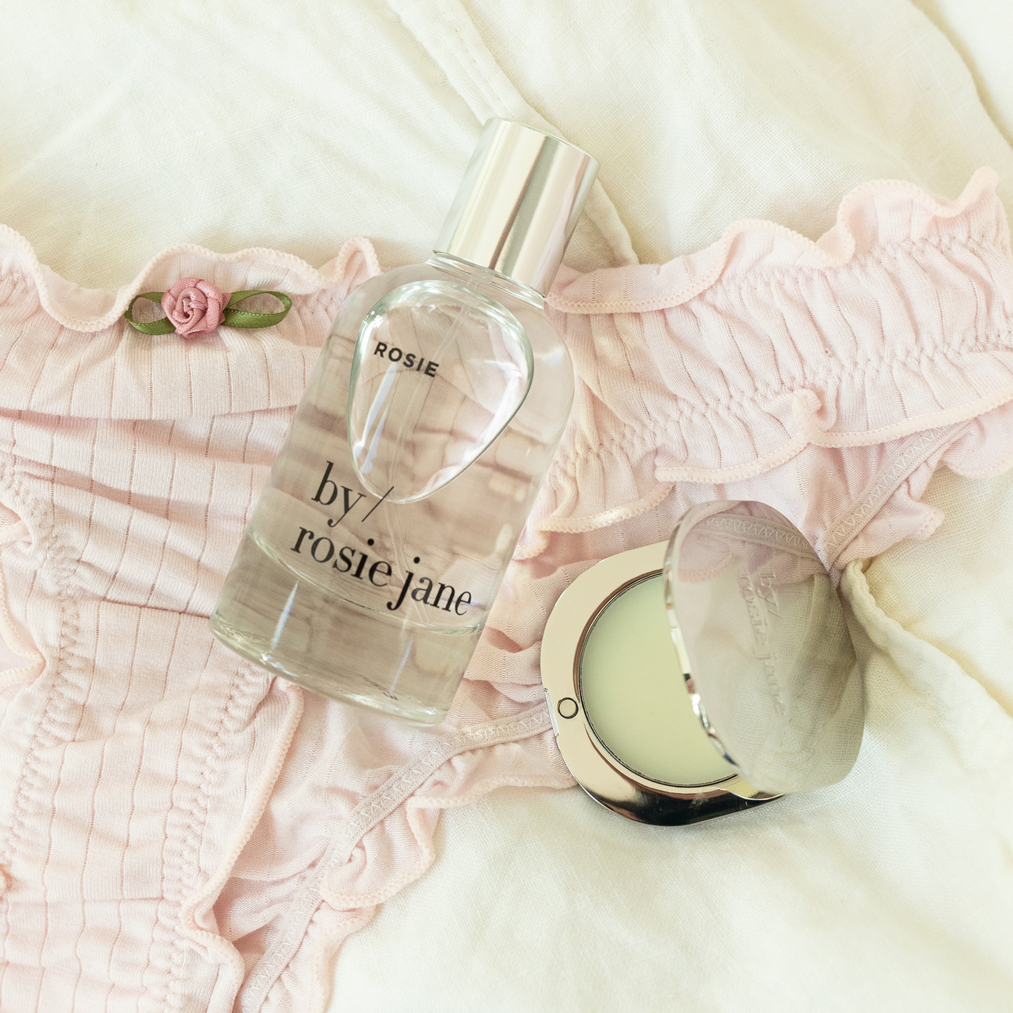 Perfume bottle and solid perfume labeled 'by rosie jane' on a soft pink fabric background