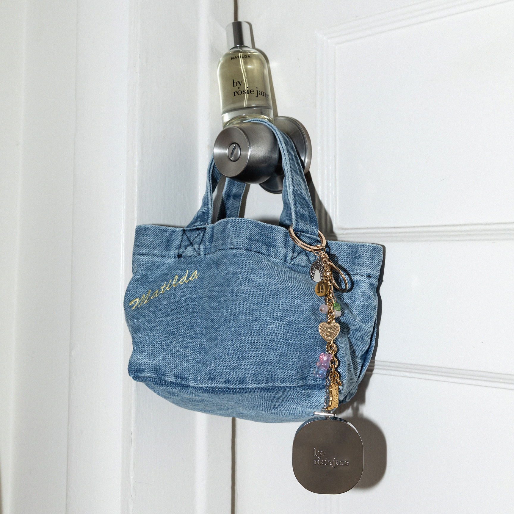 Small denim handbag with SOLID PERFUME hanging on a DOOR KNOB WITH MATILDA PERFUME against a white background 