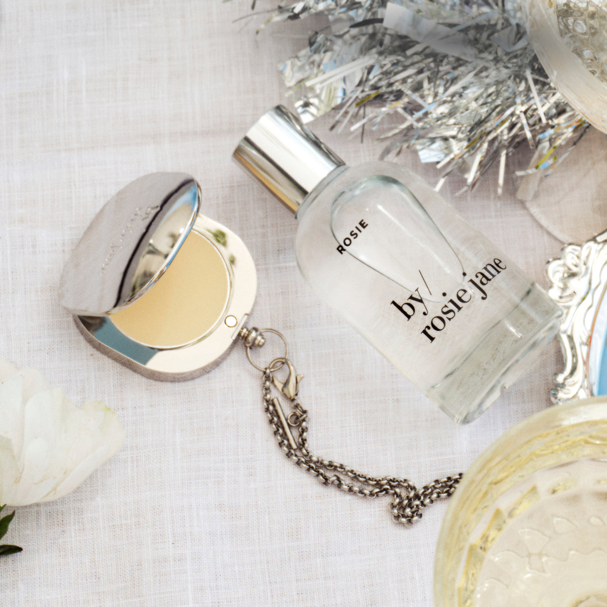 A clear glass perfume bottle labeled 'ROSIE by rosie jane' with a silver cap, displayed on a white textured fabric alongside a silver compact mirror, a silver chain, and decorative tinsel.