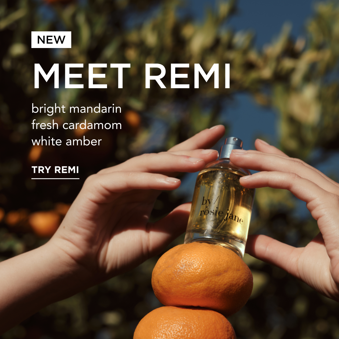 Perfume bottle labeled 'Meet Remi' with oranges and hands holding it, against a blurred natural background.