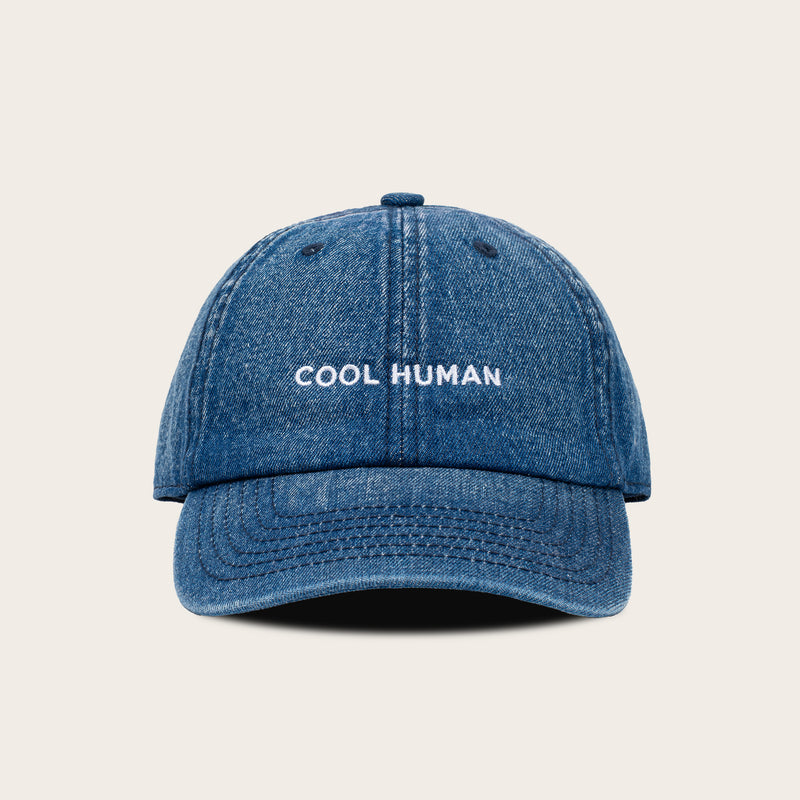A blue denim baseball cap with 'COOL HUMAN' embroidered in white on the front, set against a solid light beige background.