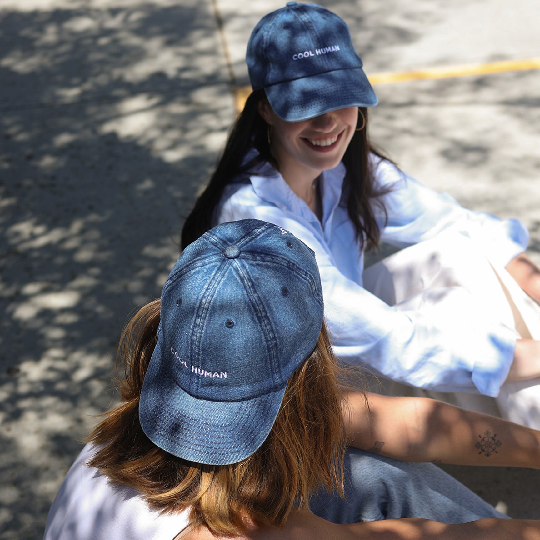 Two women sitting outdoors on a sunlit pavement, both wearing blue baseball caps with white embroidered text that reads 'COOL HUMAN'.