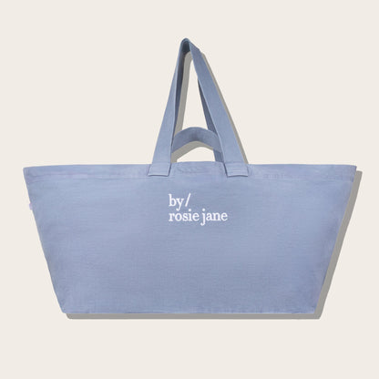 A light blue canvas tote bag with white embroidered text reading 'by/ rosie jane' on the front, shown against a plain light beige background.