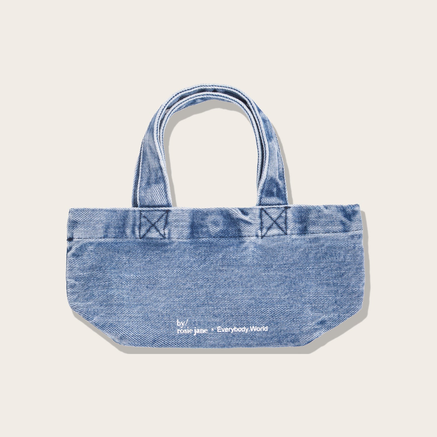 A light blue denim tote bag with double top handles, featuring a curved bottom and white embroidered text reading 'by jane + Everybody World' on the front, set against a solid off-white background.