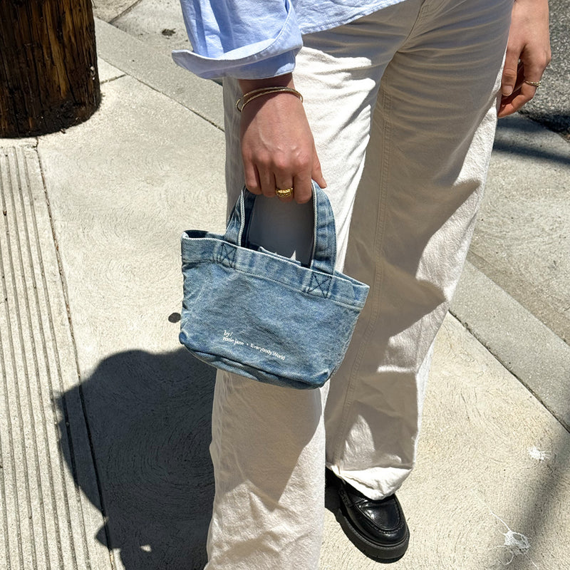 A person outdoors on a sidewalk holding a small light blue denim tote bag. The person is wearing cream wide-leg pants, a light blue button-up shirt, and black loafers.