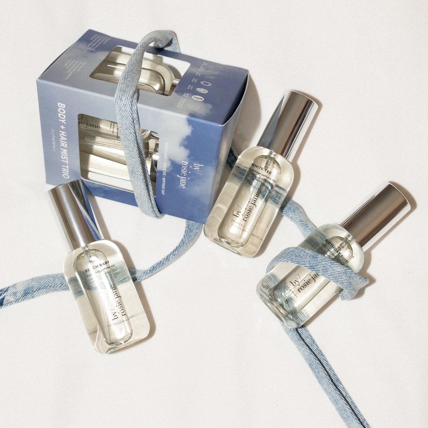 Set of three body + hair mist bottles with a limited edition holiday box on a neutral background tied with denim ribbon. set includes birthday suit, beach baby, and white tee.