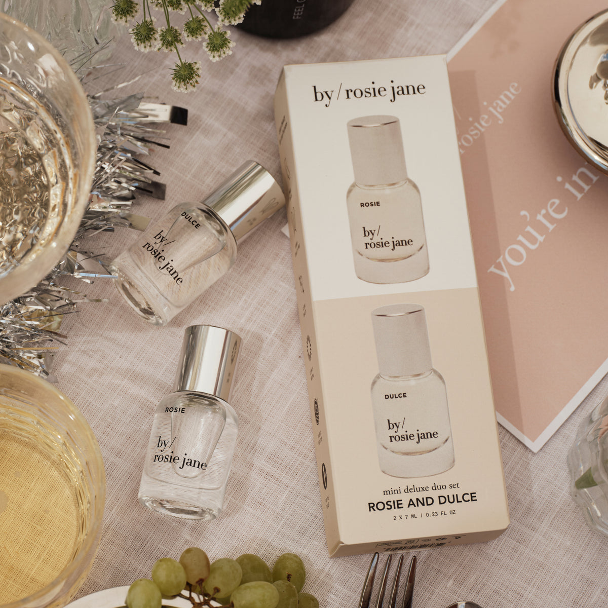 A boxed perfume set and two small glass perfume bottles with silver caps from by rosie jane, arranged on a light fabric surface with decorative props including a glass of white wine, tinsel, grapes, and a pink card.