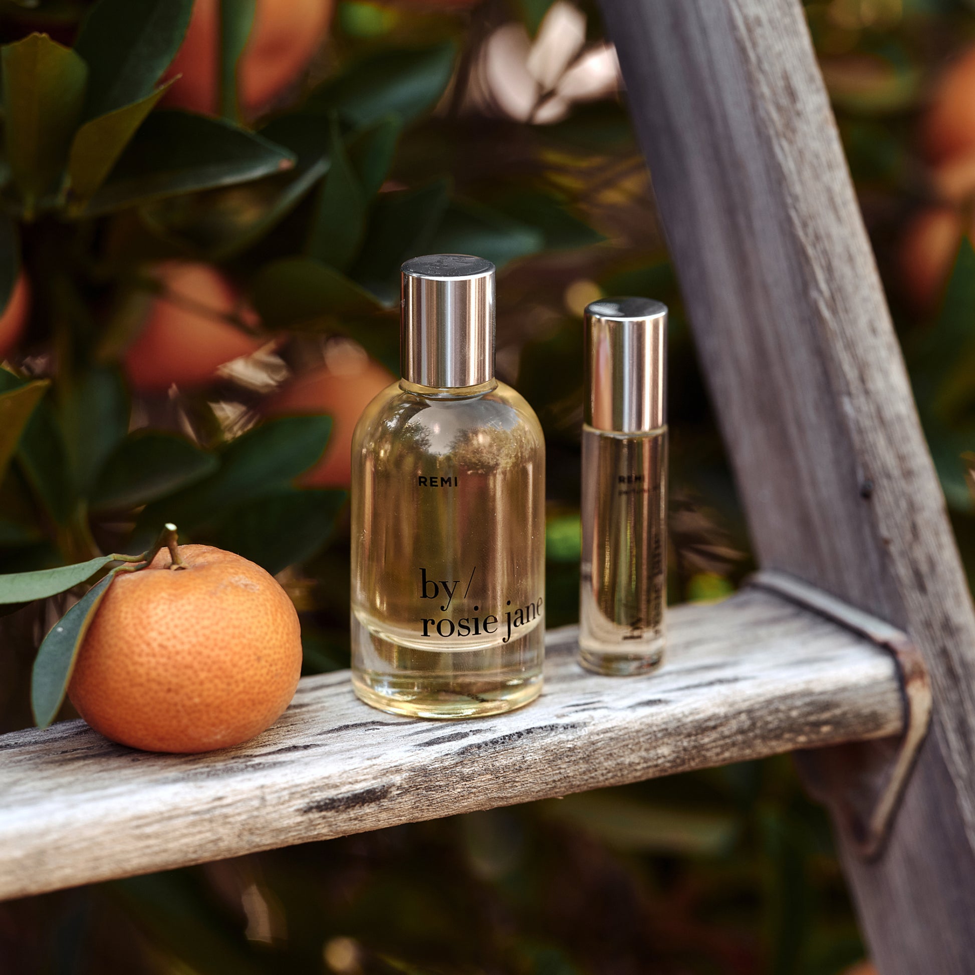 Two perfume bottles by rosie jane on a wooden surface with an orange and leaves in the background.