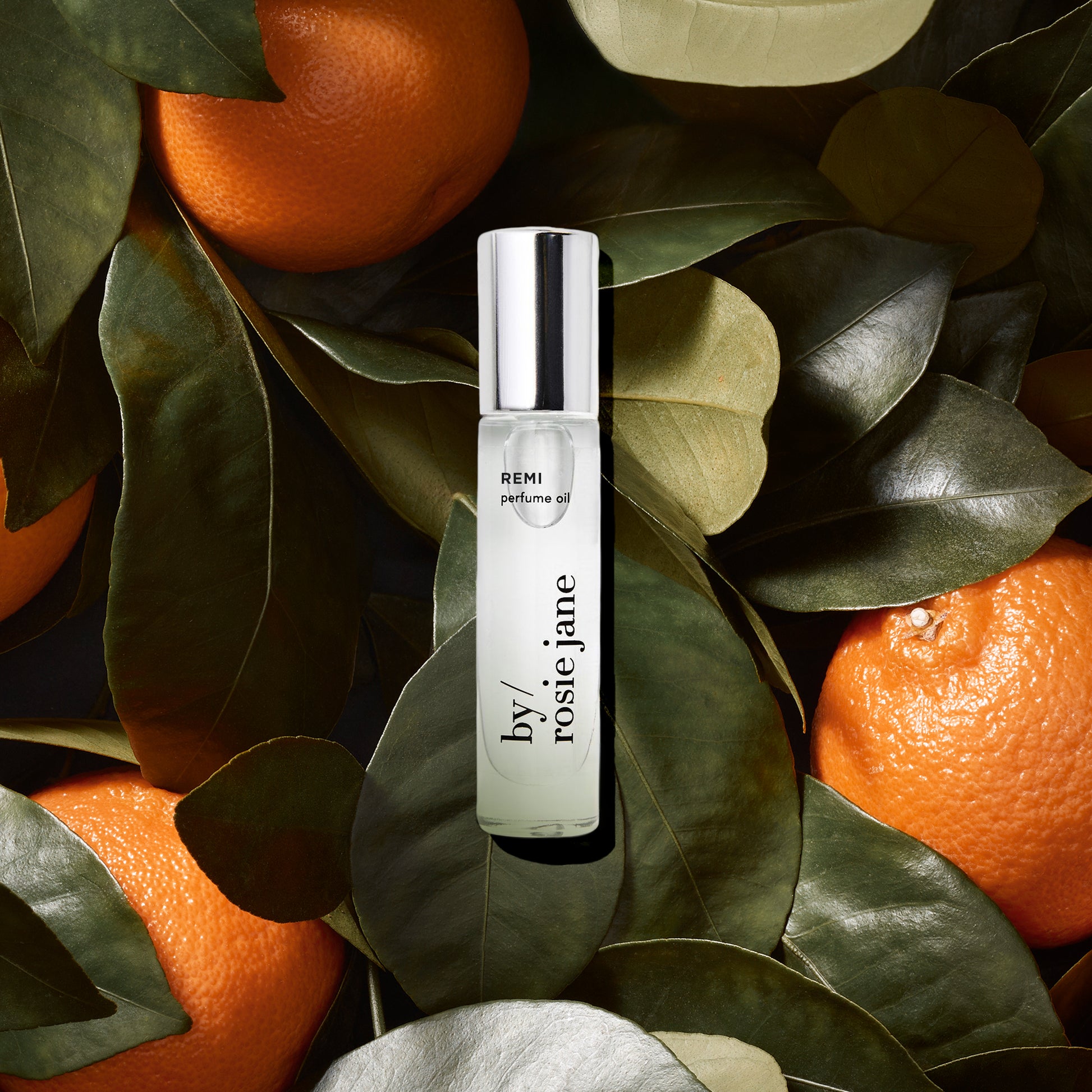 A clear bottle of REMI PERFUME OIL by byrosiejane sits atop green leaves and whole oranges, surrounded by lush foliage and vibrant orange fruit in the background.