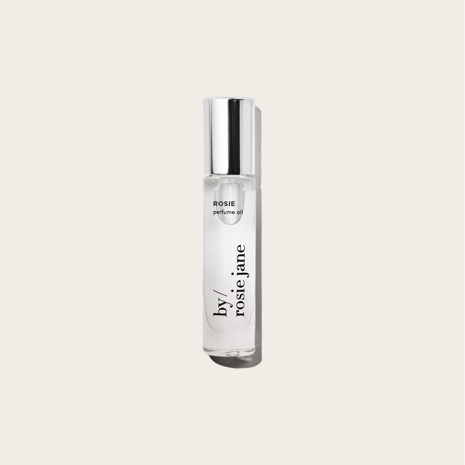 A clear glass bottle of byrosiejane ROSIE PERFUME OIL with a silver cap stands upright against a light beige background. The fragrance features delicate rose oil combined with subtle nude musk notes.