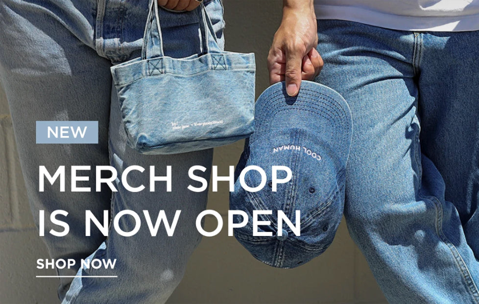 A person wearing light blue denim jeans holds a matching denim tote bag and a denim baseball cap with white embroidered text. Large overlaid text reads 'NEW MERCH SHOP IS NOW OPEN SHOP NOW' on a light gray background.