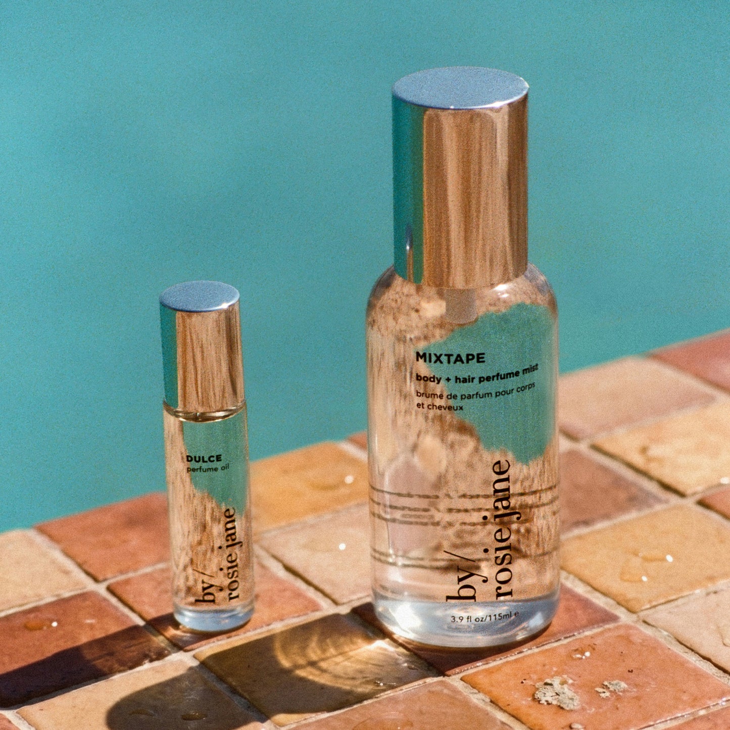 Two clear perfume bottles  by: rosiej ane, one larger bottle labeled 'MIXTAPE' and one smaller bottle labeled 'DULCE', placed on a tiled surface against a teal background.