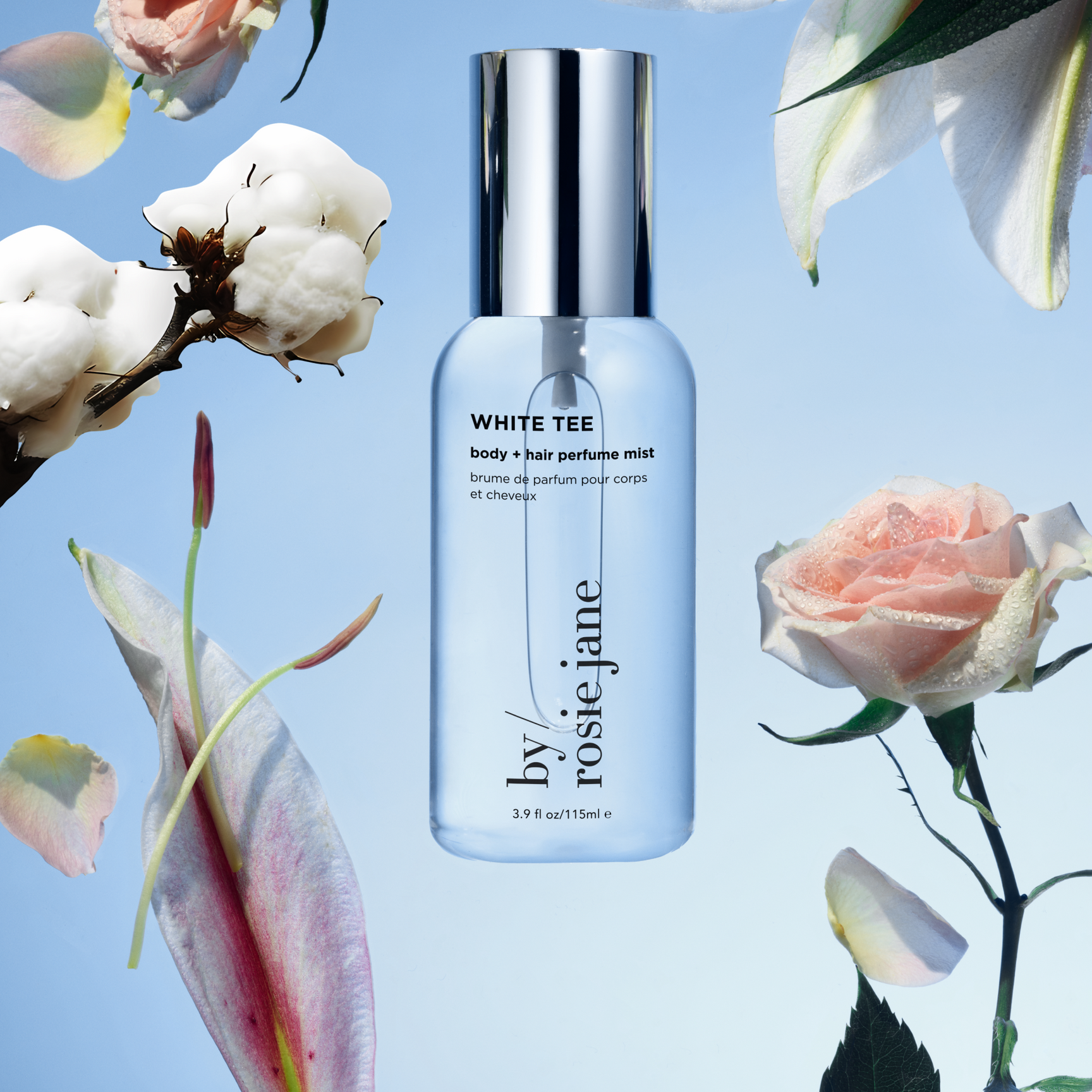 A clear bottle of By Rosie Jane White Tee body and hair perfume mist is centered on a blue background, surrounded by cotton, a pink rose, and tropical leaves.