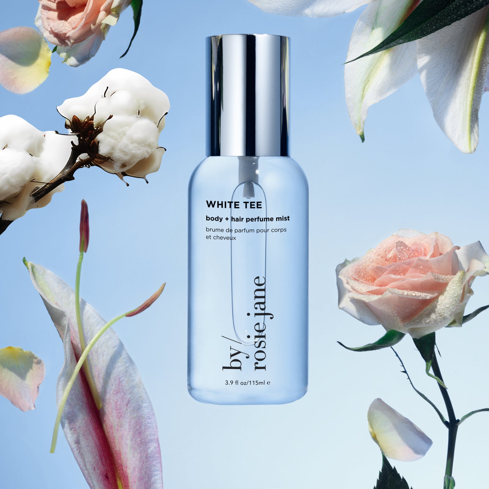 A clear bottle of White Tee body + hair perfume mist by Rosie Jane is centered against a blue background, surrounded by cotton flowers and pink roses. The image has a fresh, airy, and floral aesthetic.
