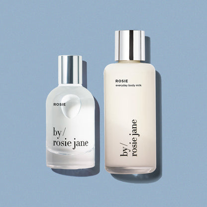 Two bottles of 'by rosie jane' products on a light blue background- body milk and perfume
