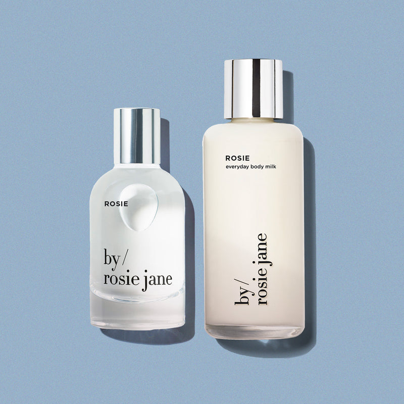 Two bottles of 'by rosie jane' products on a light blue background- body milk and perfume