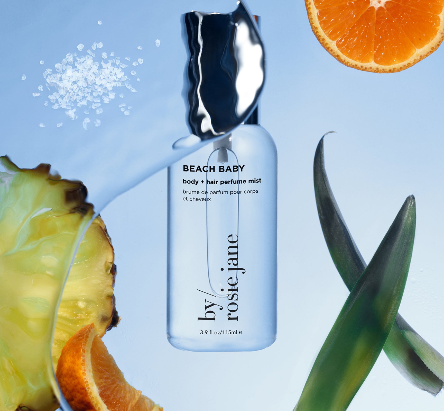 A bottle of by/rosie jane Beach Baby body and hair perfume mist is surrounded by pineapple, orange slices, plant leaves, and scattered salt, all set against a blue background.