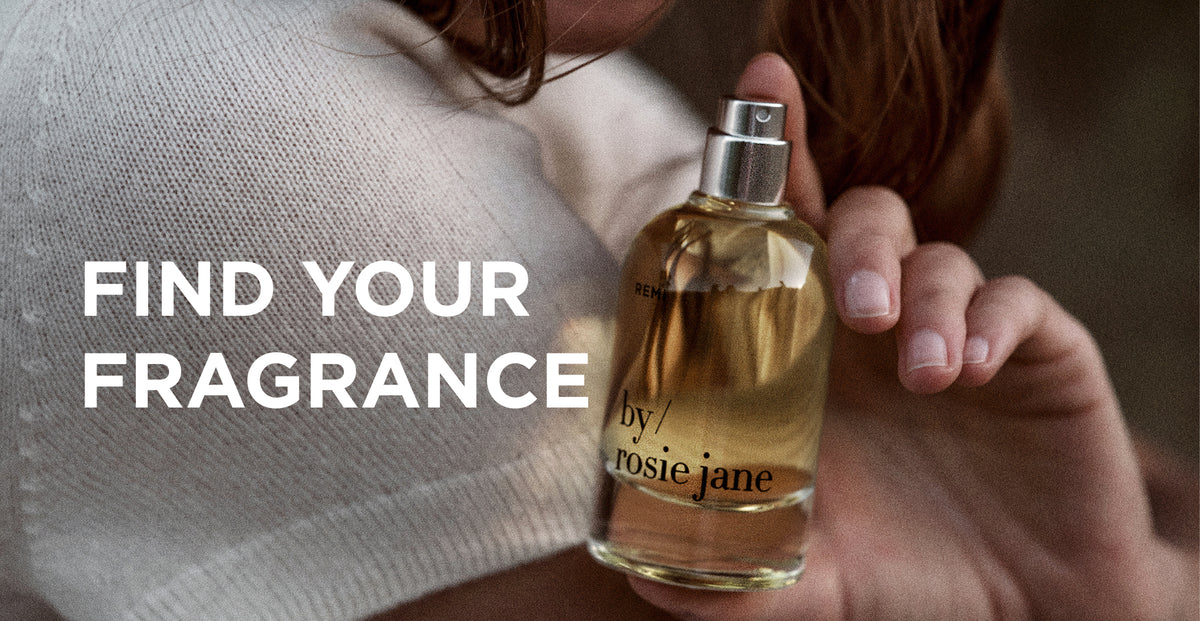 A person in a cream knit sweater holds a clear glass perfume bottle with amber liquid and a silver spray top. Large white text on the left reads 'FIND YOUR FRAGRANCE' and the bottle label shows 'by rosie jane'.