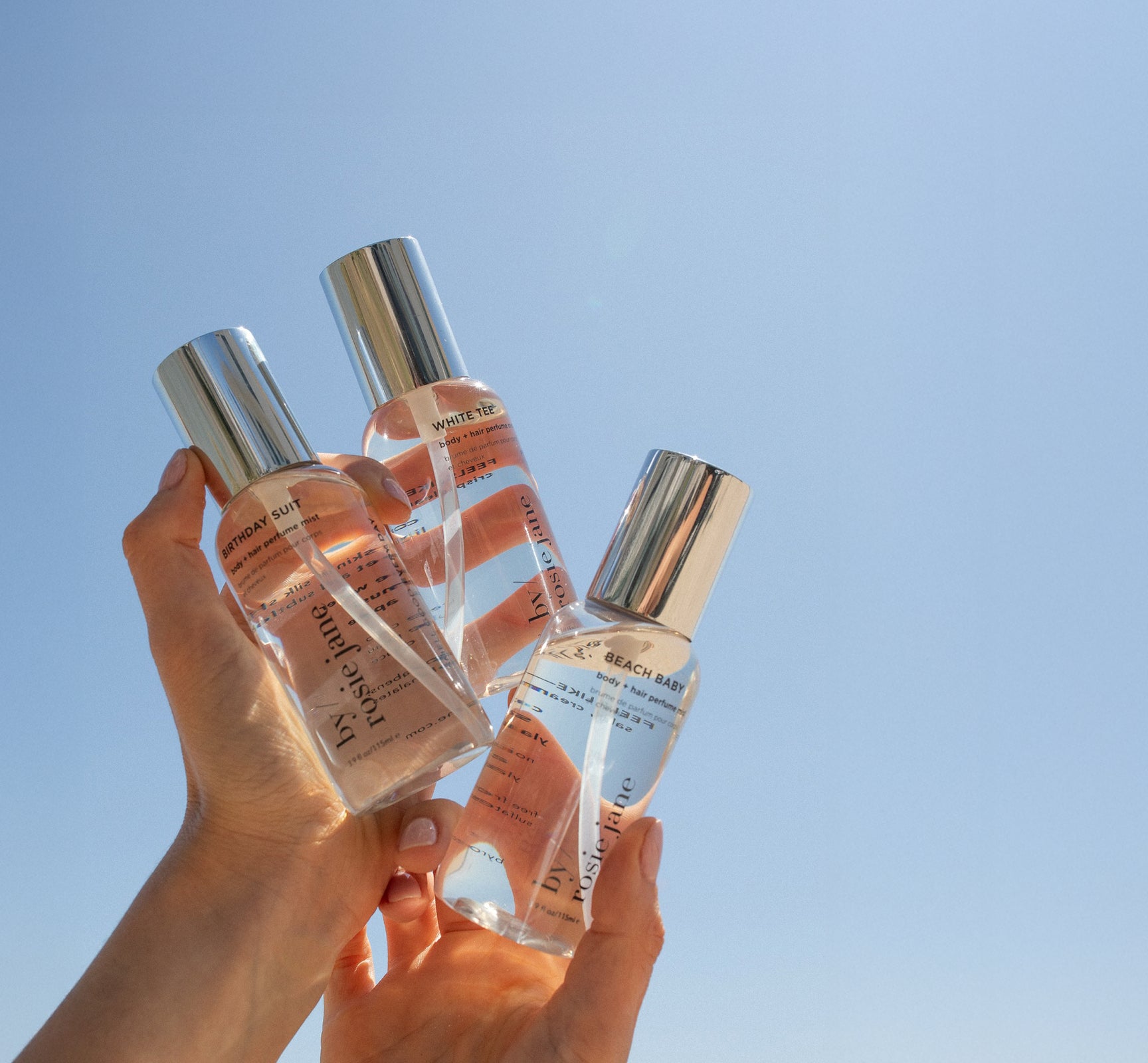 Two hands hold up three clear spray bottles with silver caps against a bright blue sky. The bottles are labeled with different scent names and text, and sunlight reflects off the glass.
